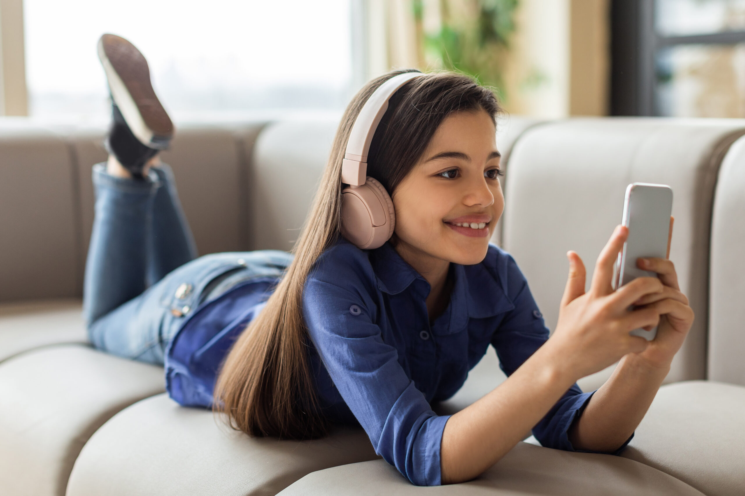 Smiling Adolescent Girl Using Mobile Phone Wearing Wireless Headphones Indoor