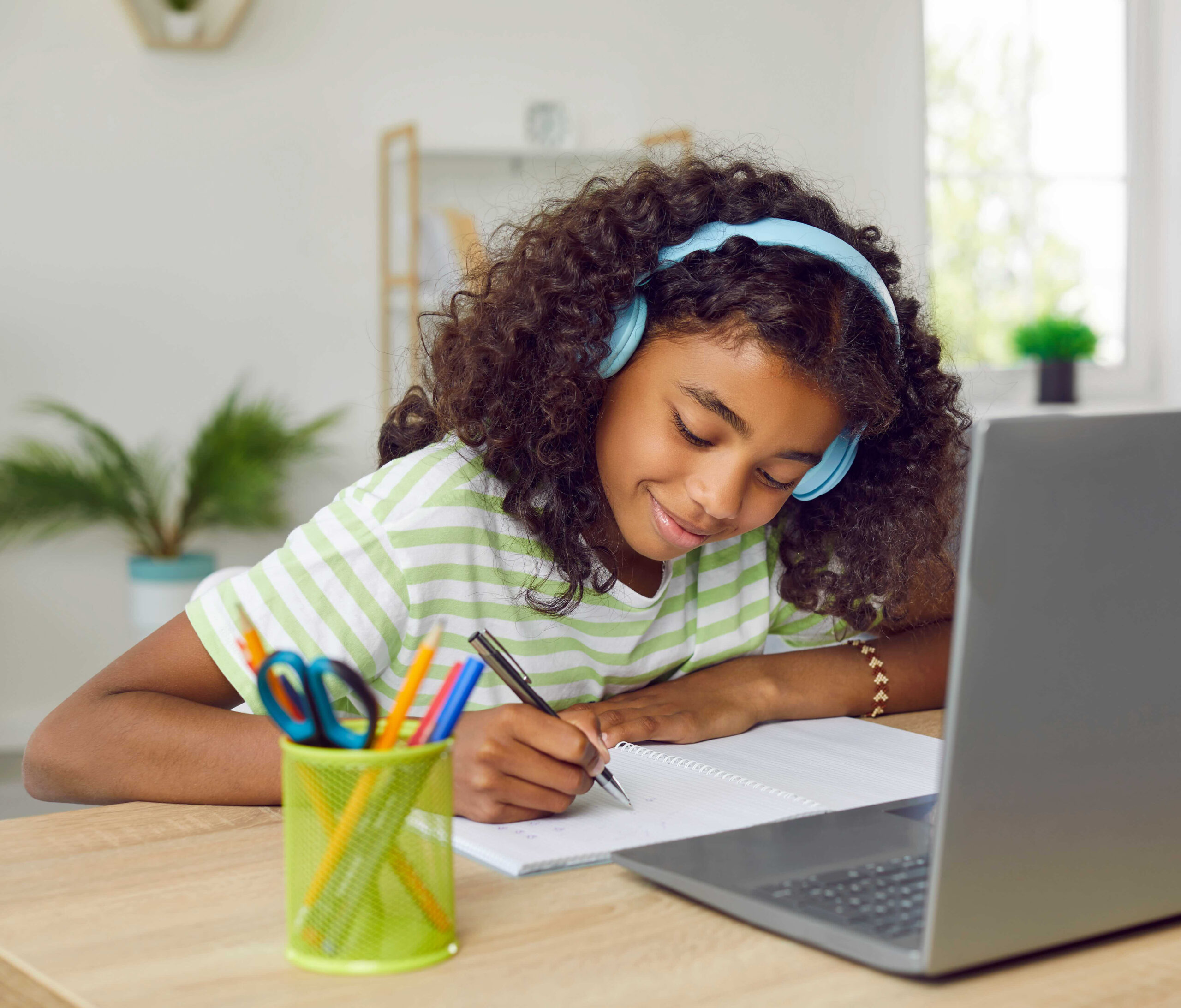 Happy school child sitting at desk with laptop, having online class and writing in notebook