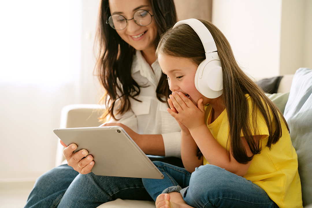 Smiling mother in glasses holding digital tablet in front of little girl daughter wearing headphones looking excited