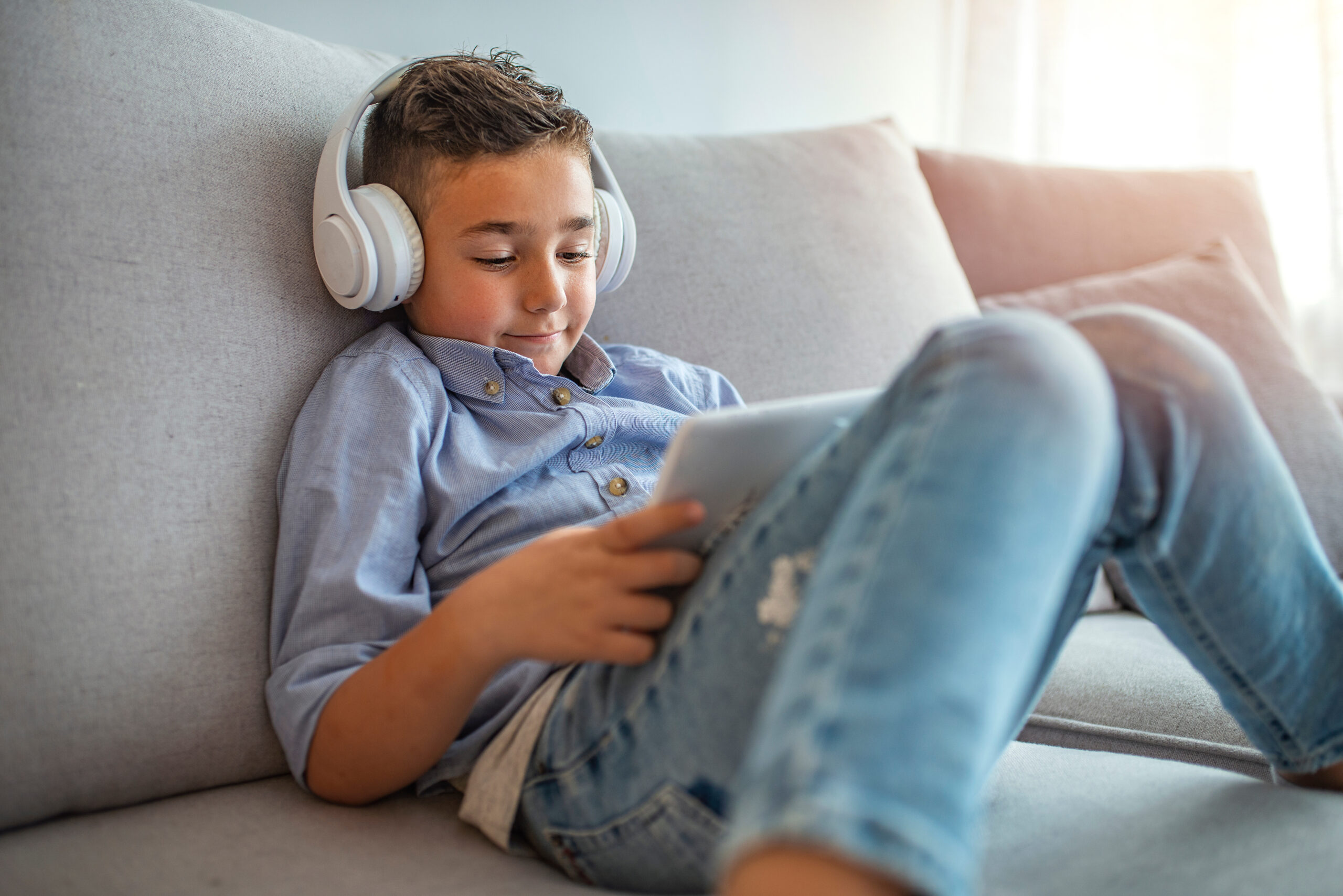 Smiling boy in big white headphones watching videos on tablet computer. Little boy in headphones is using a digital tablet and smiling. Concentrated kid playing with digital tablet